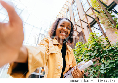 African woman student smiling to camera, making selfie photo and holding a stack of books. Concept of happiness and accomplishment, as the woman is proud of her studies or her current situation. 116339174