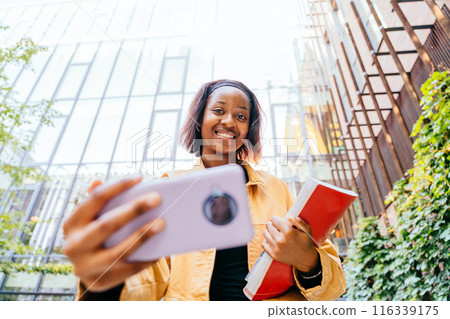 A young African student woman making selfie photo with cell phone and stack of books. She is smiling and she is happy A young African student woman making selfie photo with cell phone and stack of books. She is smiling and she is happy 116339175
