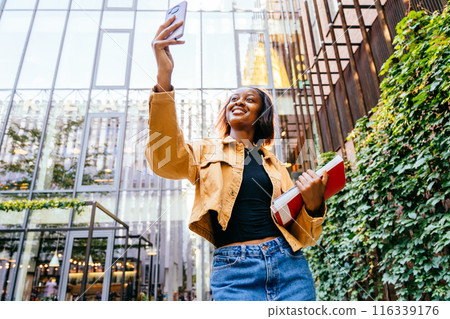 Portrait of black ethnic girl in good mood wearing casually walking in college or university talking on phone, having video call back to school. Cheerful African college girl outdoors on modern campus 116339176
