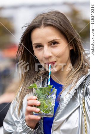 A woman is happily enjoying a refreshing cold drink outdoors on a sunny day, savoring the moment 116340318