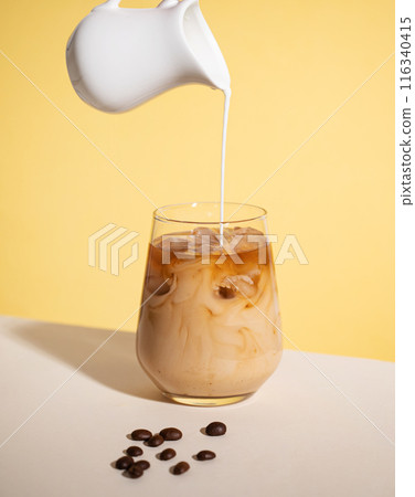 Iced coffee with poured cream in a glass with ice cubes and grains on a yellow background.  116340415