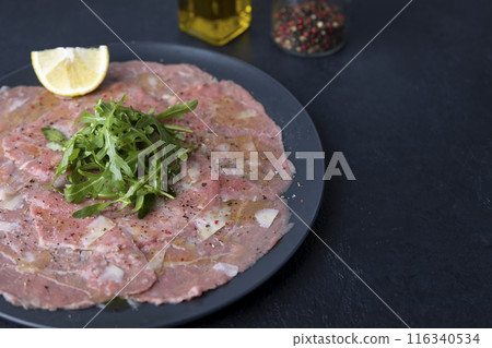 Marbled beef carpaccio with arugula, parmesan cheese, olive oil and lemon. A traditional classic appetizer made from thinly sliced raw meat. Close-up, selective focus, black background. 116340534