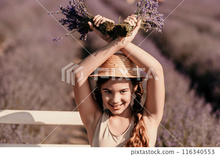 girl is holding a bunch of lavender purple flowers in her hands and wearing a straw hat. She is smiling and she is enjoying herself. The scene is set in a field of lavender, which adds to the peaceful 116340553