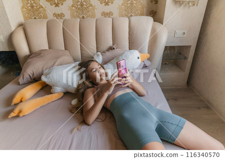 A girl is laying on a bed with a pink phone in her hand. She is looking at the phone and she is focused on something on the screen. 116340570