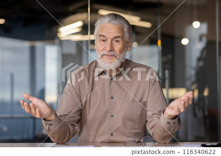 Senior businessman with gray beard in a meeting gestures with his hands, expressing ideas in a modern office. The professional looks engaged thoughtful, surrounded by contemporary interior design. Senior businessman with gray beard in a meeting gestures with his hands, expressing ideas in a modern office. The professional looks engaged thoughtful, surrounded by contemporary interior design. 116340622