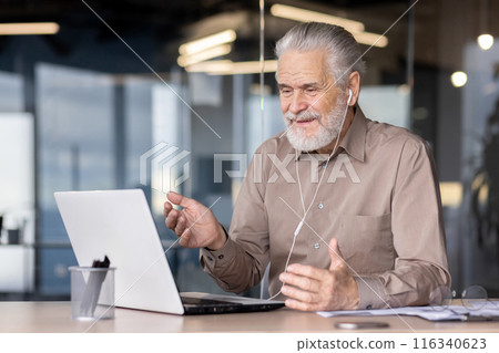 An elderly man is engaged in a video call, wearing headphones and smiling, while working on a laptop at a modern office. The image conveys communication, modern technology, professional engagement. 116340623