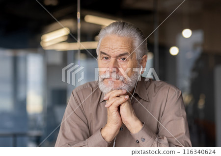 Close-up of a thoughtful senior man with gray beard and headphones, sitting in an office environment. The image captures a moment of reflection and concentration. 116340624