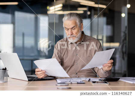 Older male professional analyzing paperwork while working on a laptop in a contemporary office setting. The man is focusing on the details of the documents, reflecting concentration and diligence. Older male professional analyzing paperwork while working on a laptop in a contemporary office setting. The man is focusing on the details of the documents, reflecting concentration and diligence. 116340627