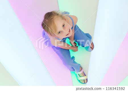 Happy 7-year-old girl in stylish denim suit looking at camera, sitting on rainbow illuminated stairs Happy 7-year-old girl in stylish denim suit looking at camera, sitting on rainbow illuminated stairs 116341592