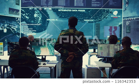 Army officer examining satellite surveillance footage on a big screen for reconnaissance, gathering intelligence to ensure national safety. Secret service federal agency works on defense. Camera B. 116342353