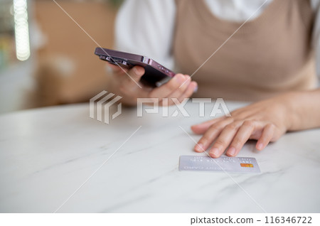 A close-up image of a woman sitting at a table indoor with her smartphone and credit card. 116346722