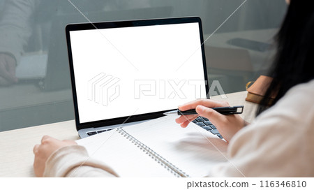 A woman working remotely from a coffee shop, using a laptop while taking notes in her notebook. 116346810