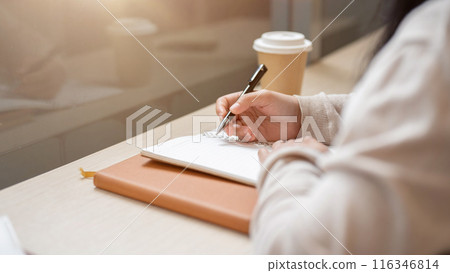 A close-up image of a woman holding a pen, writing in a book at a table by the window in the cafe. 116346814