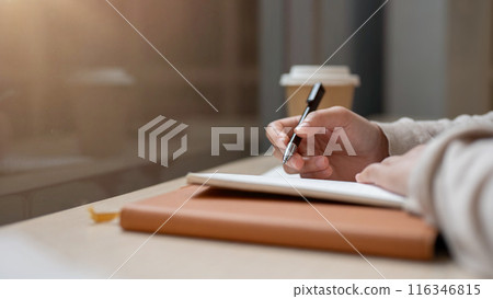 A close-up image of a woman holding a pen, writing in a book at a table by the window in the cafe. A close-up image of a woman holding a pen, writing in a book at a table by the window in the cafe. 116346815