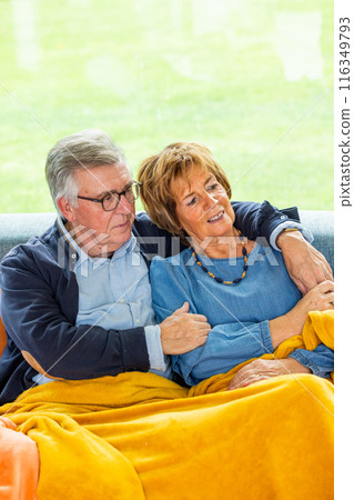 Elderly couple cuddling under a blanket on the sofa, happily together in their home Elderly couple cuddling under a blanket on the sofa, happily together in their home 116349793