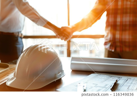 Two people shaking hands in front of a hard hat 116349836