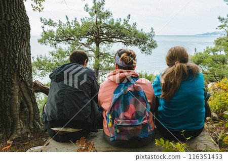 Three people sit on a wooden bench overlooking a scenic view of the sea, surrounded by trees. Each person is wearing a jacket or hoodie, and one has a colorful backpack. Three people sit on a wooden bench overlooking a scenic view of the sea, surrounded by trees. Each person is wearing a jacket or hoodie, and one has a colorful backpack. 116351453