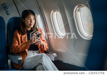 Woman seated on airplane, using smartphone, wearing orange blazer and white pants, looking out window, in-flight travel. 116351756