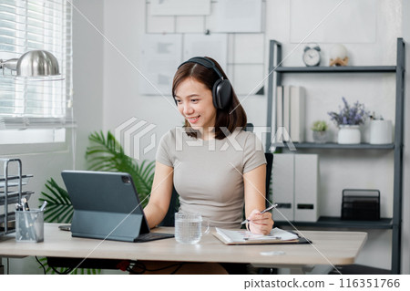 A young woman wearing headphones works from home using a tablet in a modern home office. She is smiling and taking notes. A young woman wearing headphones works from home using a tablet in a modern home office. She is smiling and taking notes. 116351766