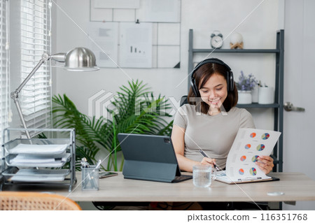 Young woman in home office wearing headphones, analyzing charts and graphs on tablet and paper, modern workspace. 116351768