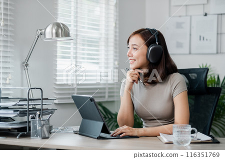 A young woman wearing headphones works from home, using a tablet and laptop in a modern, bright home office setting. 116351769