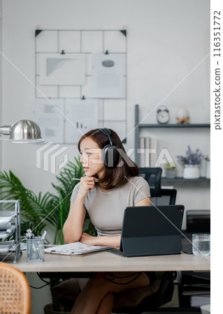 A young woman wearing headphones, working from home at a modern desk setup with a tablet and office supplies. A young woman wearing headphones, working from home at a modern desk setup with a tablet and office supplies. 116351772