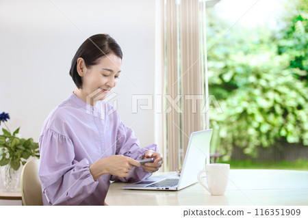 A middle-aged woman looking at a computer while using her smartphone in the living room 116351909