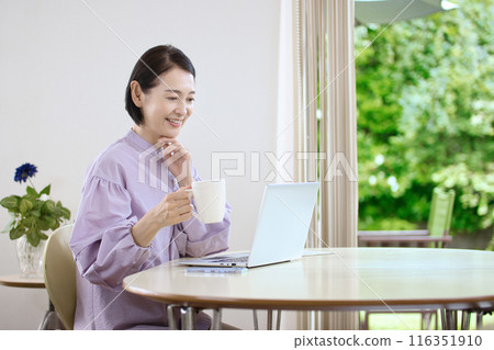 A middle-aged woman drinking coffee and looking at a computer in the living room 116351910