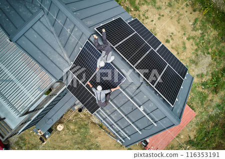 Mounters building photovoltaic solar module station on roof of house. Men electricians in helmets installing solar panel system outdoors. Concept of alternative and renewable energy. Aerial view. 116353191