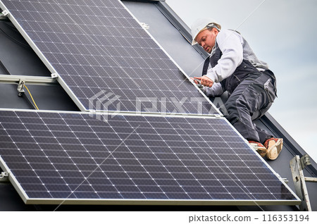 Man engineer mounting photovoltaic solar panels on roof of house. Technician in helmet installing solar module system with help of hex key. Concept of alternative, renewable energy. Man engineer mounting photovoltaic solar panels on roof of house. Technician in helmet installing solar module system with help of hex key. Concept of alternative, renewable energy. 116353194