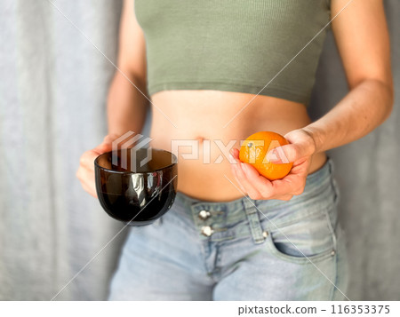 Close up of woman in jeans and green top holding of hands fresh orange and black cup of coffee. Concept of healthy eating and lifestyle. On neutral background with selective focus. 116353375