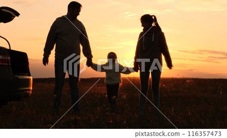 Family holding hands at sunset, silhouettes near car. Joint recreation in nature. Mother father daughter in orange setting sun in field. Family picnic road trip, parents kid enjoy each other's company 116357473