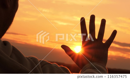 Silhouette of hand at orange sunset. Man stretches hand towards sun, warming palm in summer rays. Unity with nature, enjoyment of trip. Guy gains strength, contemplating, enjoying solitude with nature Silhouette of hand at orange sunset. Man stretches hand towards sun, warming palm in summer rays. Unity with nature, enjoyment of trip. Guy gains strength, contemplating, enjoying solitude with nature 116357475
