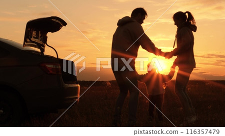Silhouettes of family dancing at sunset near car. Mom dad kid holding hands jumping spinning around laughing smiling. Family of travelers celebrating road trip, picnic in nature. Active rest at nature 116357479