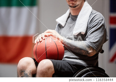 Cropped shot of tattooed basketball player with disability using wheelchair and holding ball in lap with dramatic lighting in indoor court copy space 116358444