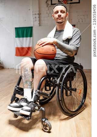 Vertical portrait of tattooed adult man with disability using wheelchair in indoor basketball court and looking at camera with dramatic light Vertical portrait of tattooed adult man with disability using wheelchair in indoor basketball court and looking at camera with dramatic light 116358456