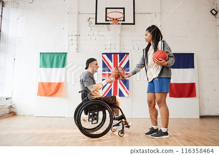 Side view portrait of young basketball player with disability using wheelchair during practice with female coach indoors copy space 116358464