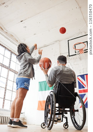 Vertical back view of sportsman using wheelchair in basketball practice with female coach throwing ball in hoop Vertical back view of sportsman using wheelchair in basketball practice with female coach throwing ball in hoop 116358470
