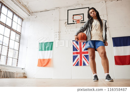 Wide angle portrait of young Black woman as female basketball player holding ball and looking at camera in indoor court with international flags in background copy space Wide angle portrait of young Black woman as female basketball player holding ball and looking at camera in indoor court with international flags in background copy space 116358487