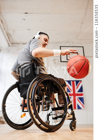 Vertical full length portrait of young man in wheelchair playing adaptive sports and dribbling ball at indoor basketball court 116358535