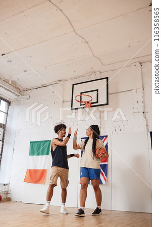Vertical full length shot of two young people man and woman playing basketball and high five in indoor court Vertical full length shot of two young people man and woman playing basketball and high five in indoor court 116358565