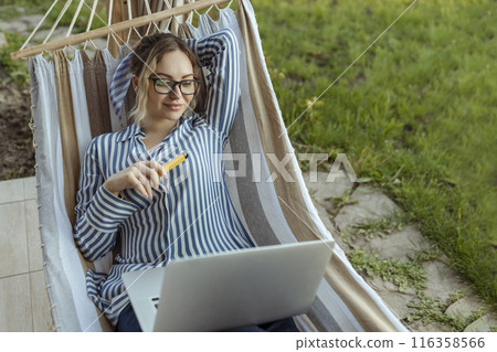 young adult girl in a hammock works on a computer outside the house and smokes an electronic cigarette young adult girl in a hammock works on a computer outside the house and smokes an electronic cigarette 116358566