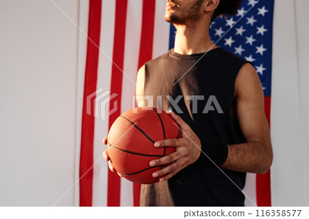 Cropped shot of unrecognizable Black young man as basketball player holding ball standing against American flag in background copy space Cropped shot of unrecognizable Black young man as basketball player holding ball standing against American flag in background copy space 116358577