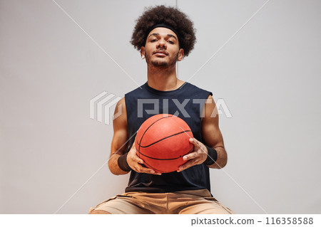 Minimal front view portrait of Black young man as basketball player holding ball and looking at camera sitting on bench against white wall copy space Minimal front view portrait of Black young man as basketball player holding ball and looking at camera sitting on bench against white wall copy space 116358588