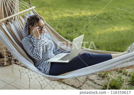 Young beautiful woman lies in a hammock with a laptop in glasses outside at home 116358617