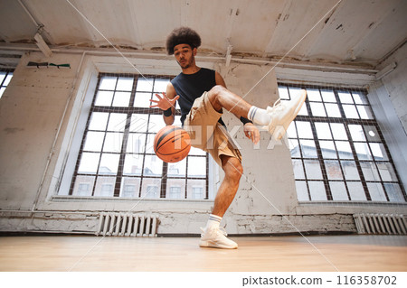 Wide angle action shot of young man as professional basketball player demonstrating tricks with ball while practicing in indoor gym copy space Wide angle action shot of young man as professional basketball player demonstrating tricks with ball while practicing in indoor gym copy space 116358702