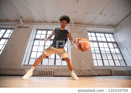 Wide angle action shot of African American basketball player dribbling ball while practicing in indoor gym copy space 116358709