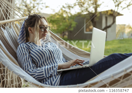 Young beautiful woman lies in a hammock with a laptop in glasses outside at home 116358747