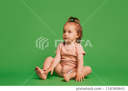 Little baby girl, child in pink bodysuit, with ponytail sitting calmly and looking isolated on green background Little baby girl, child in pink bodysuit, with ponytail sitting calmly and looking isolated on green background 116360641