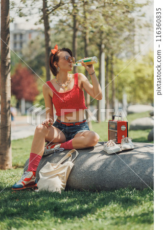 Young lady dressed vintage top and denim shorts sitting on stone, drinking lemonade, at urban park and listening music on retro radio Young lady dressed vintage top and denim shorts sitting on stone, drinking lemonade, at urban park and listening music on retro radio 116360835